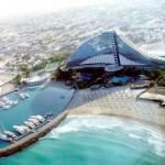 A modern waterfront building with a triangular roof by a marina and beach.