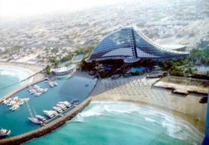 A modern waterfront building with a triangular roof by a marina and beach.