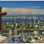 City view from a hilltop with a cross in the foreground under a clear sky.